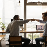 Well done, buddy. Motivated diverse young men coworkers bump fists on workplace feel excited achieve common goal. Two workers international business team members share success glad to help one another